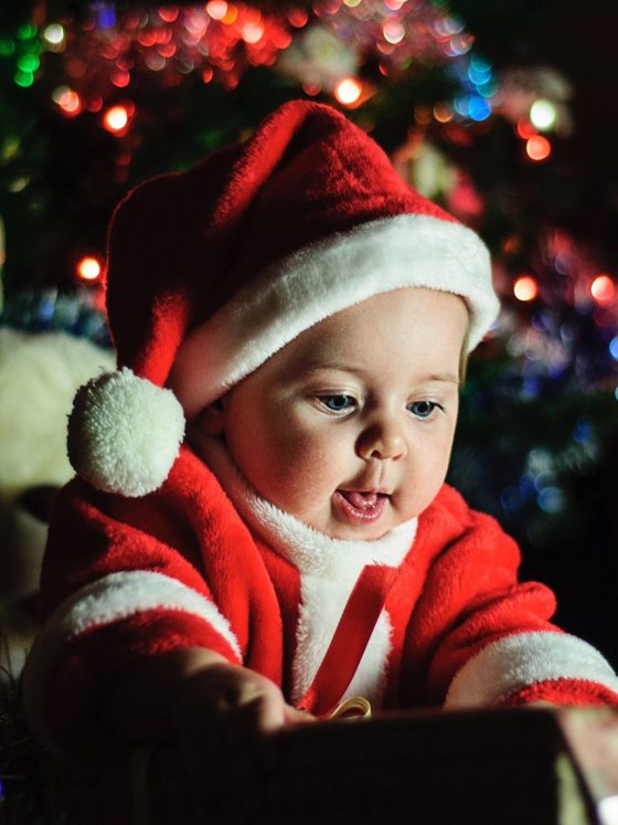 baby dressed in santa outfit opening a gift, with christmas tree in the background