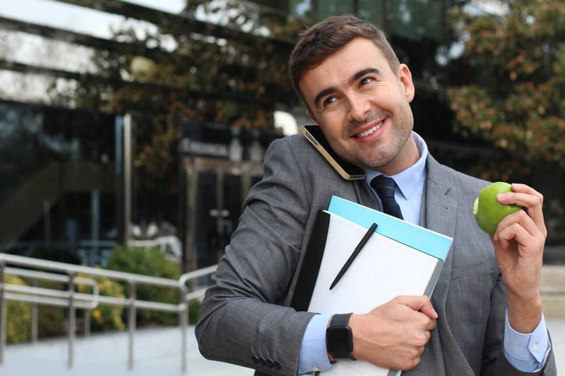 business man balancing a busy schedule, eating an apple while answering the phone, and holding work papers.